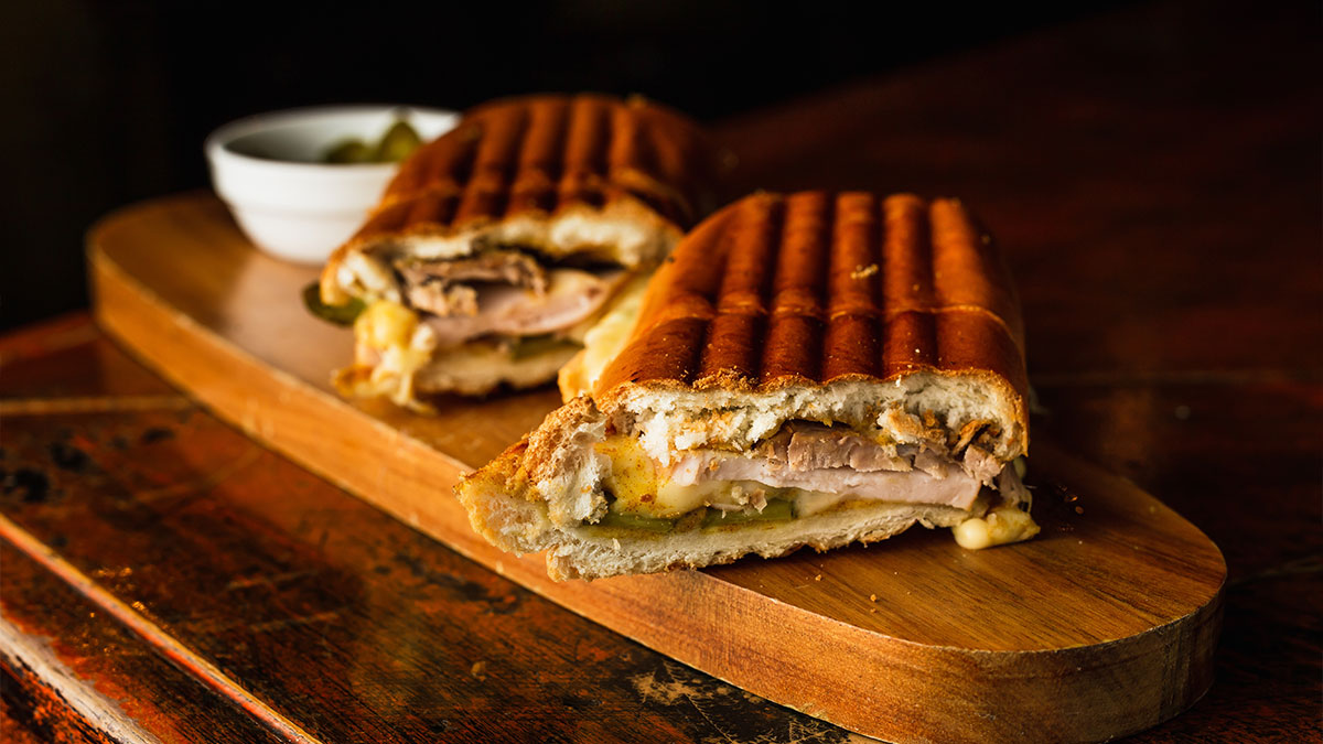 Close-up of a Cuban sandwich on a wooden cutting board, featuring layers of roast pork, ham, Swiss cheese, pickles, and mustard.